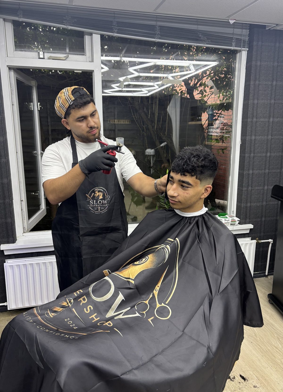 De barber aan het werk met tondeuse, in een schort met het Slow-logo, bij een klant onder een zwart-gouden cape